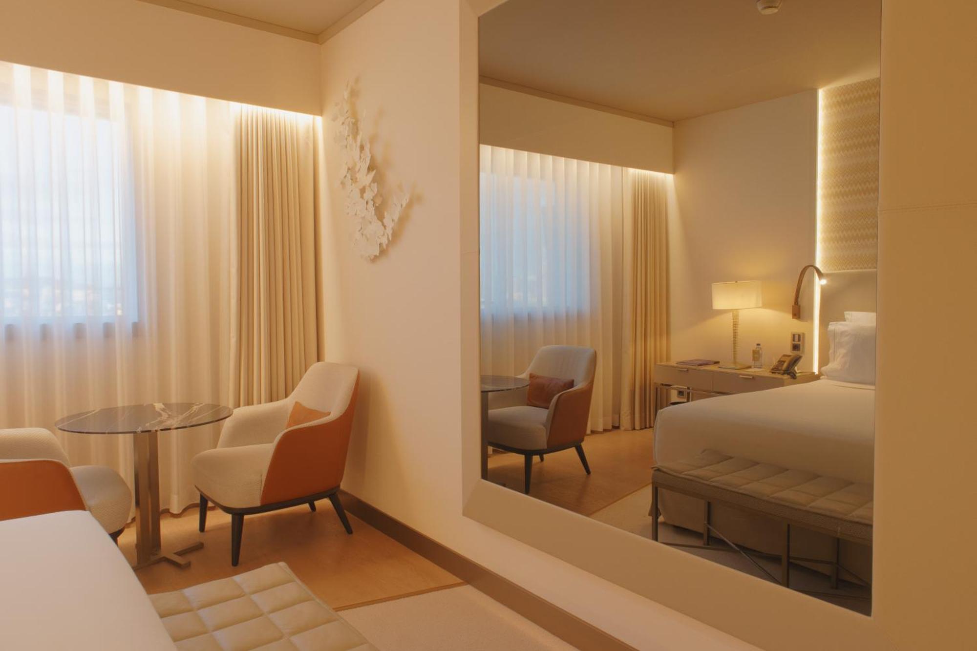 Epic Sana Marques Hotel Lissabon Exterior foto The photo showcases a modern hotel room with a minimalist and stylish design. In the foreground, there's a small round table with two chairs, one of which features an orange accent. The walls are adorned with sheer, light-colored curtains that allow 